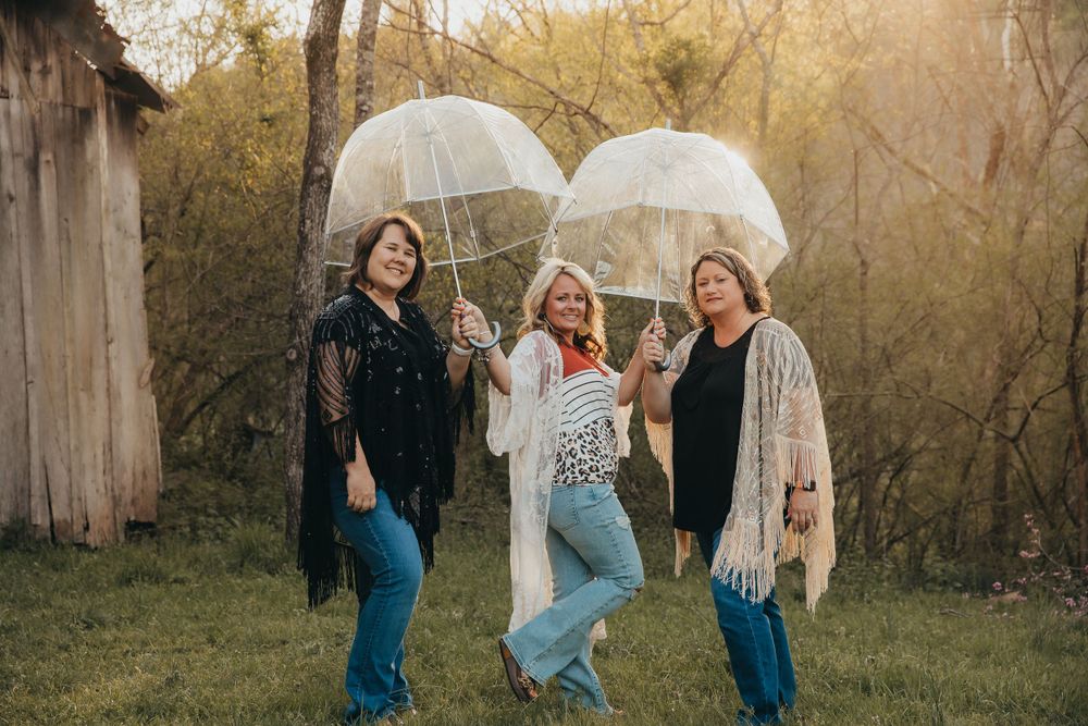 Three friends laugh together while holding clear umbrellas in a backyard during golden hour.