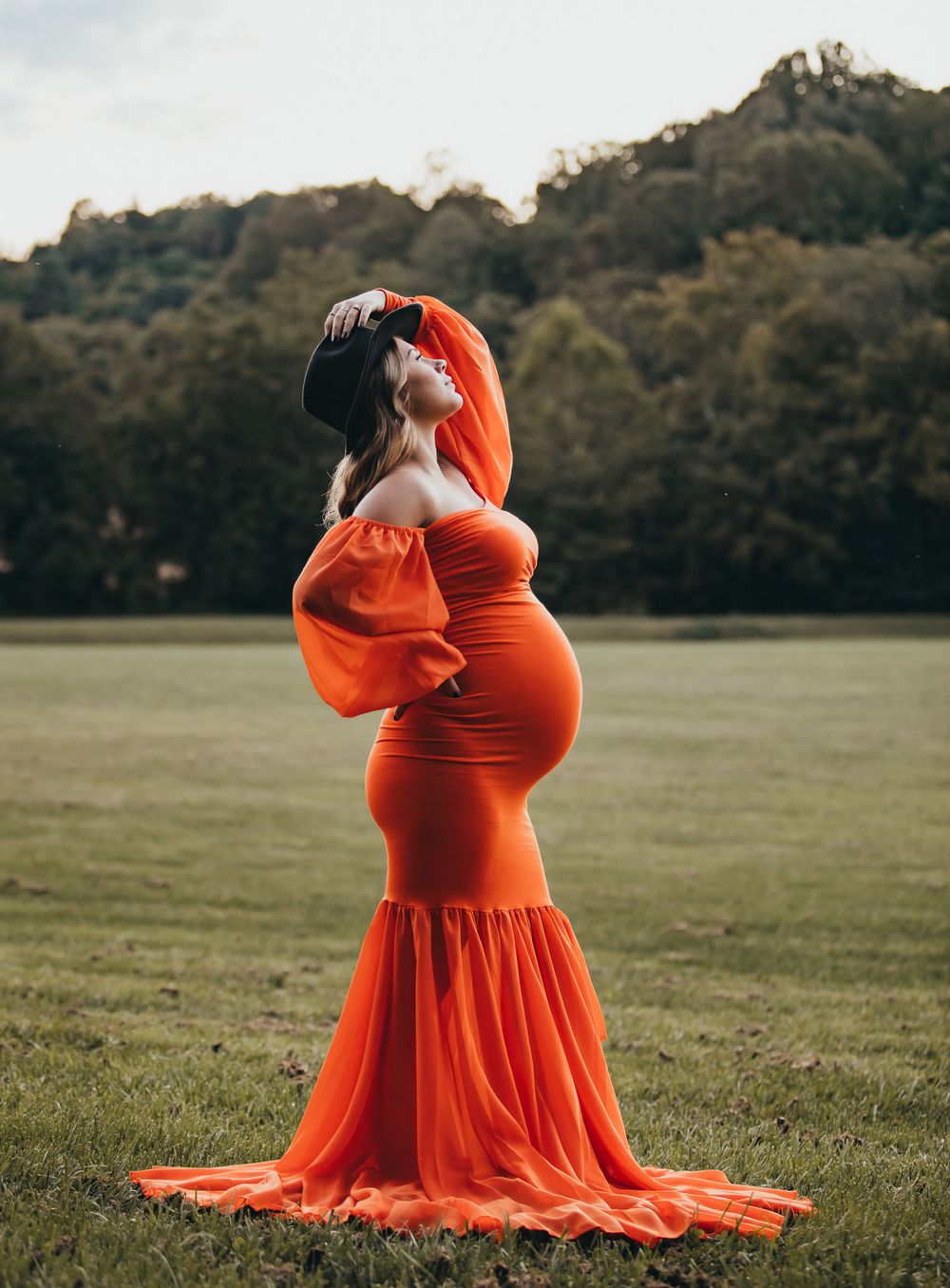 A woman in a flowing orange maternity gown poses gracefully in a grassy field at sunset.