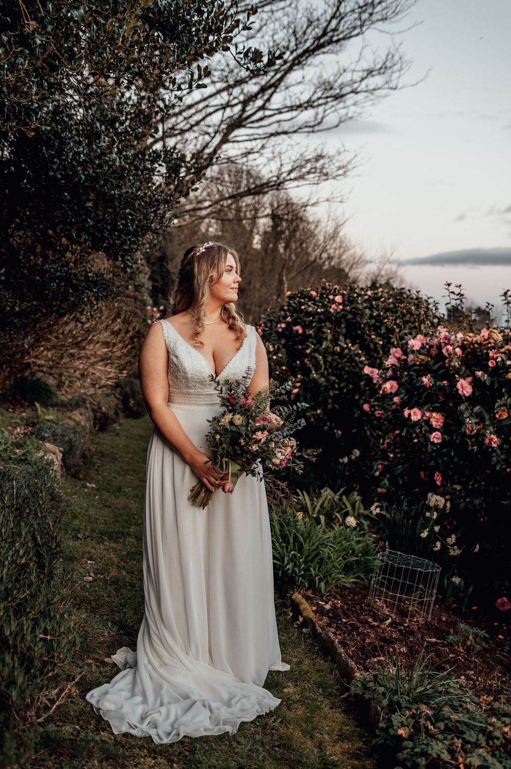 A bride in a flowing white dress stands near blooming roses at sunset, holding a bouquet in a coastal garden setting.