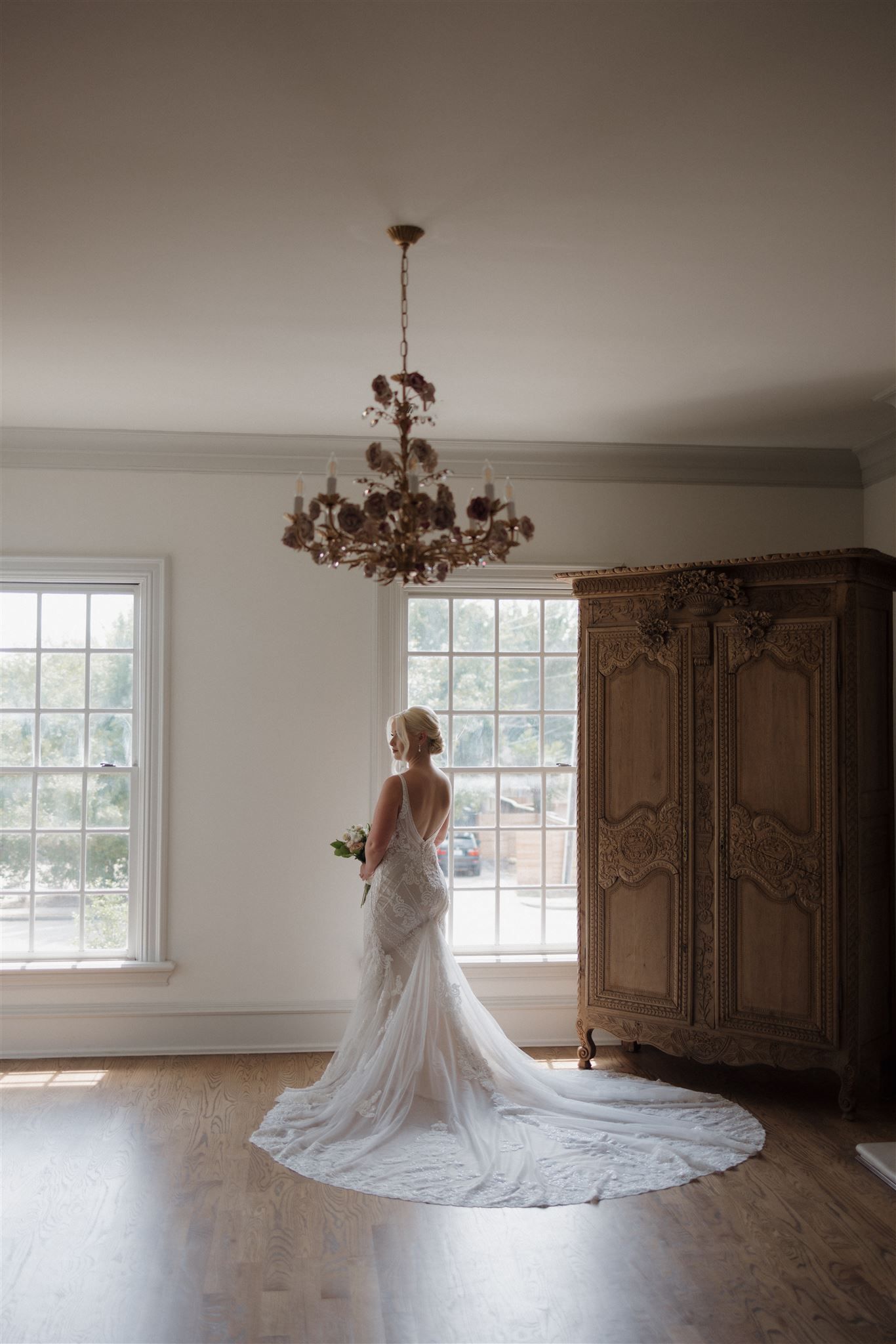 A dramatic wedding dress silhouette against windows with an antique armoire and chandelier.