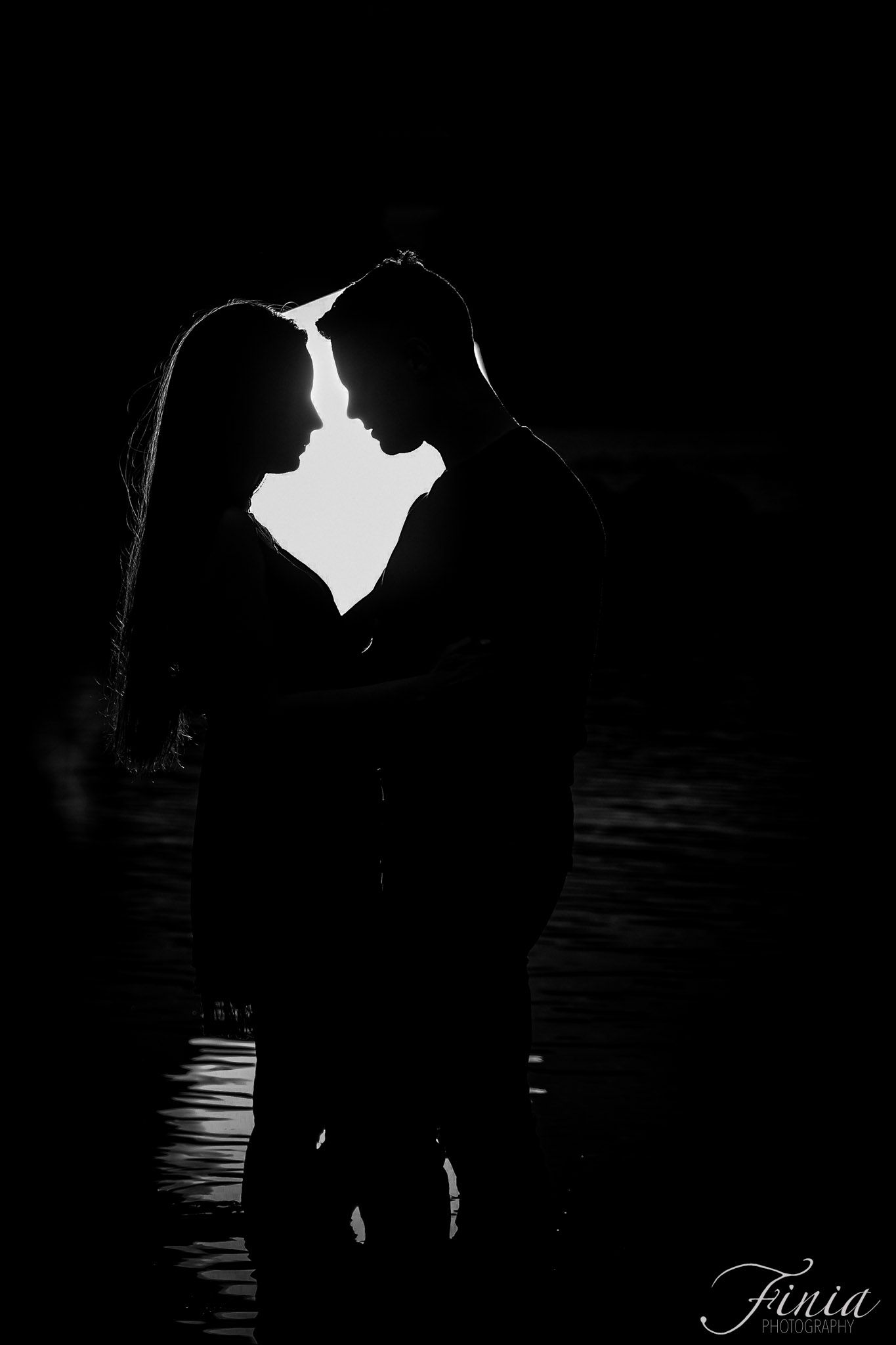 A silhouetted couple stands close together in a romantic backlit pose creating a dramatic black and white contrast.