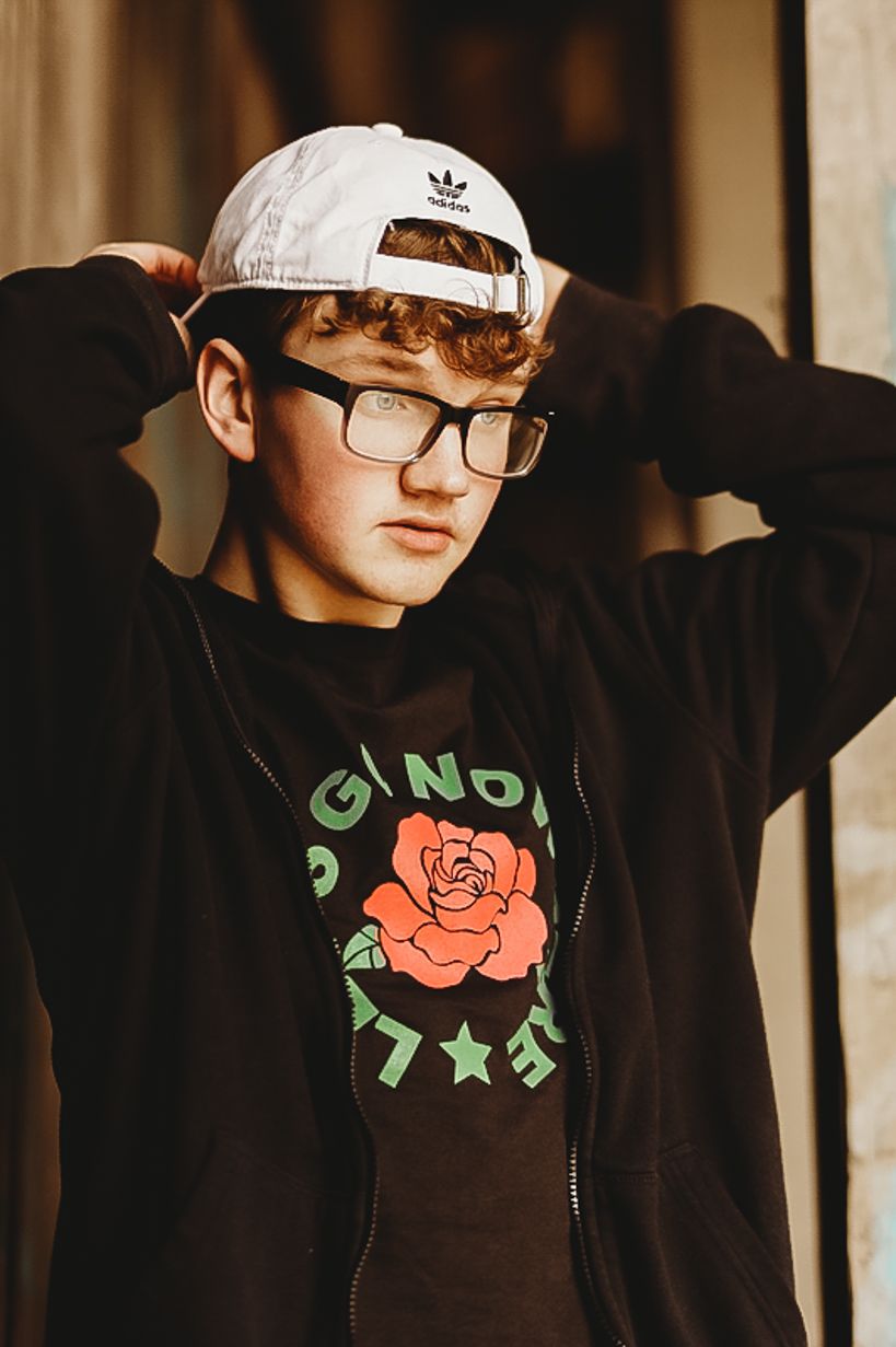 Person in black hoodie and white baseball cap adjusting hat while posing against wall.