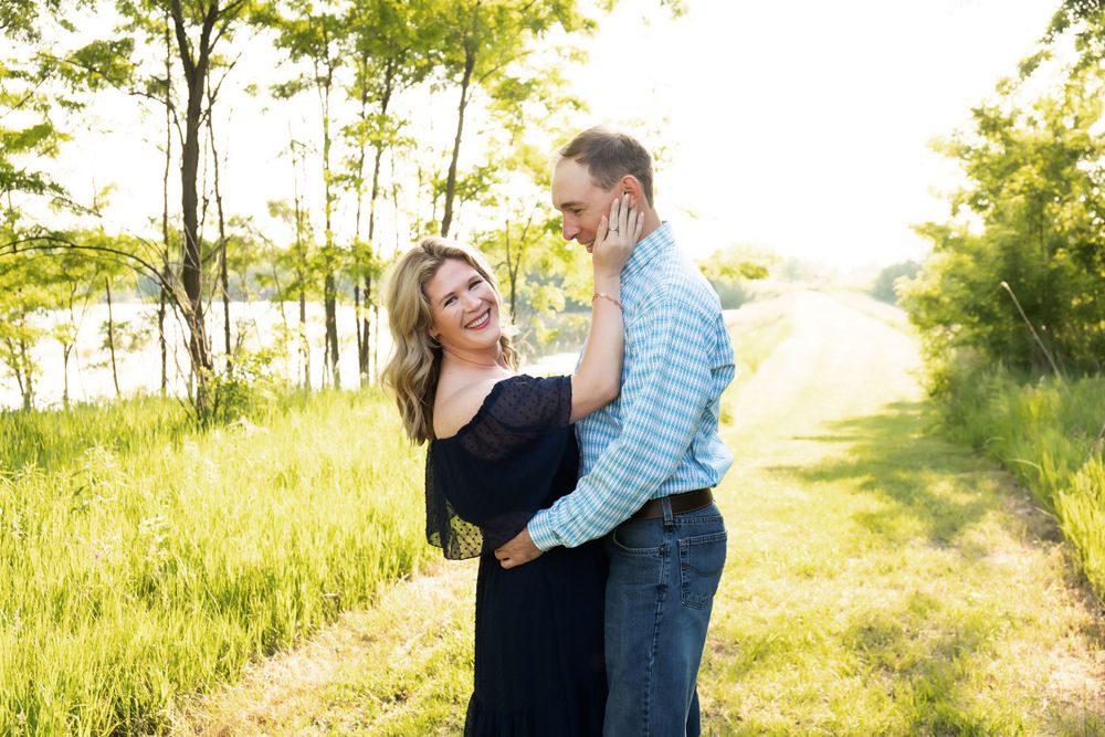A couple laughs together while standing on a grassy path with trees in the background.