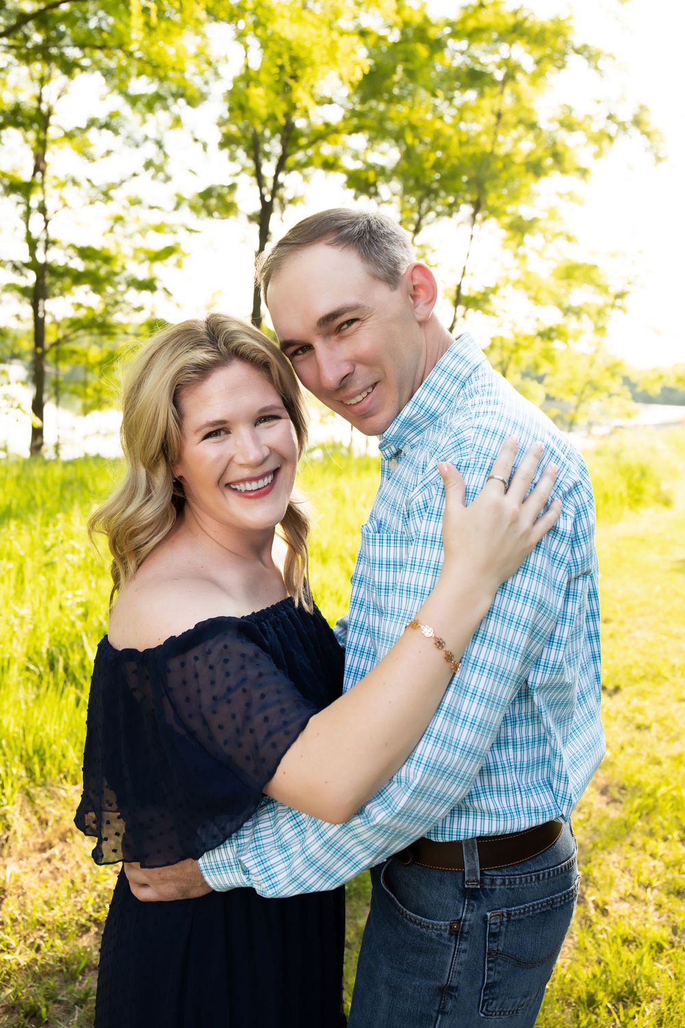 A couple in coordinating outfits smiles while embracing in a sunny meadow.