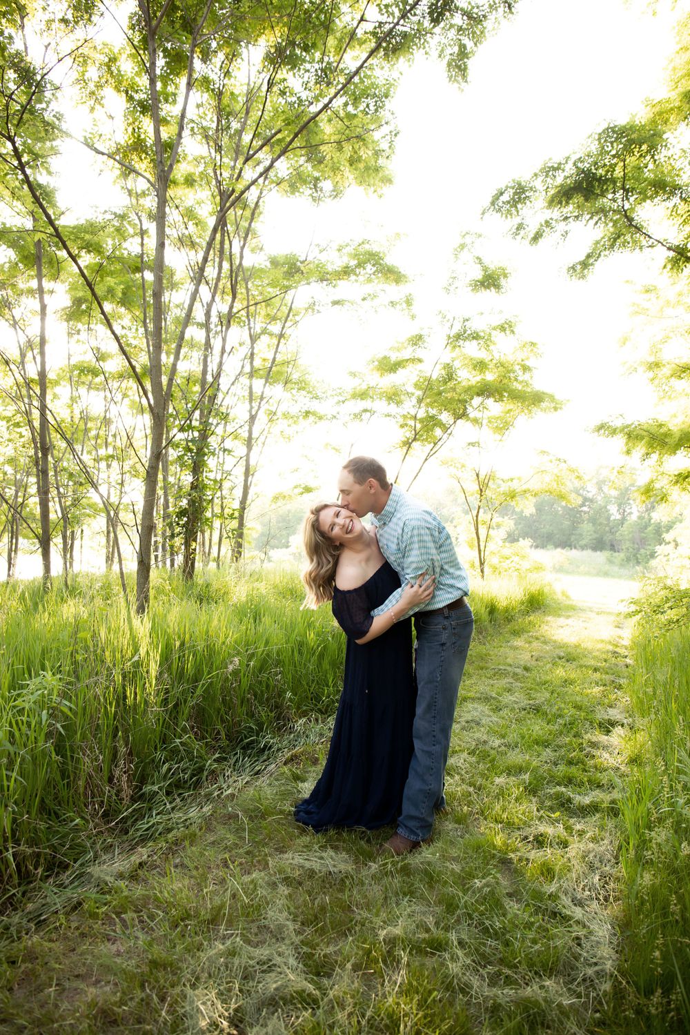 A couple embraces on a nature trail surrounded by tall trees and evening sunlight.