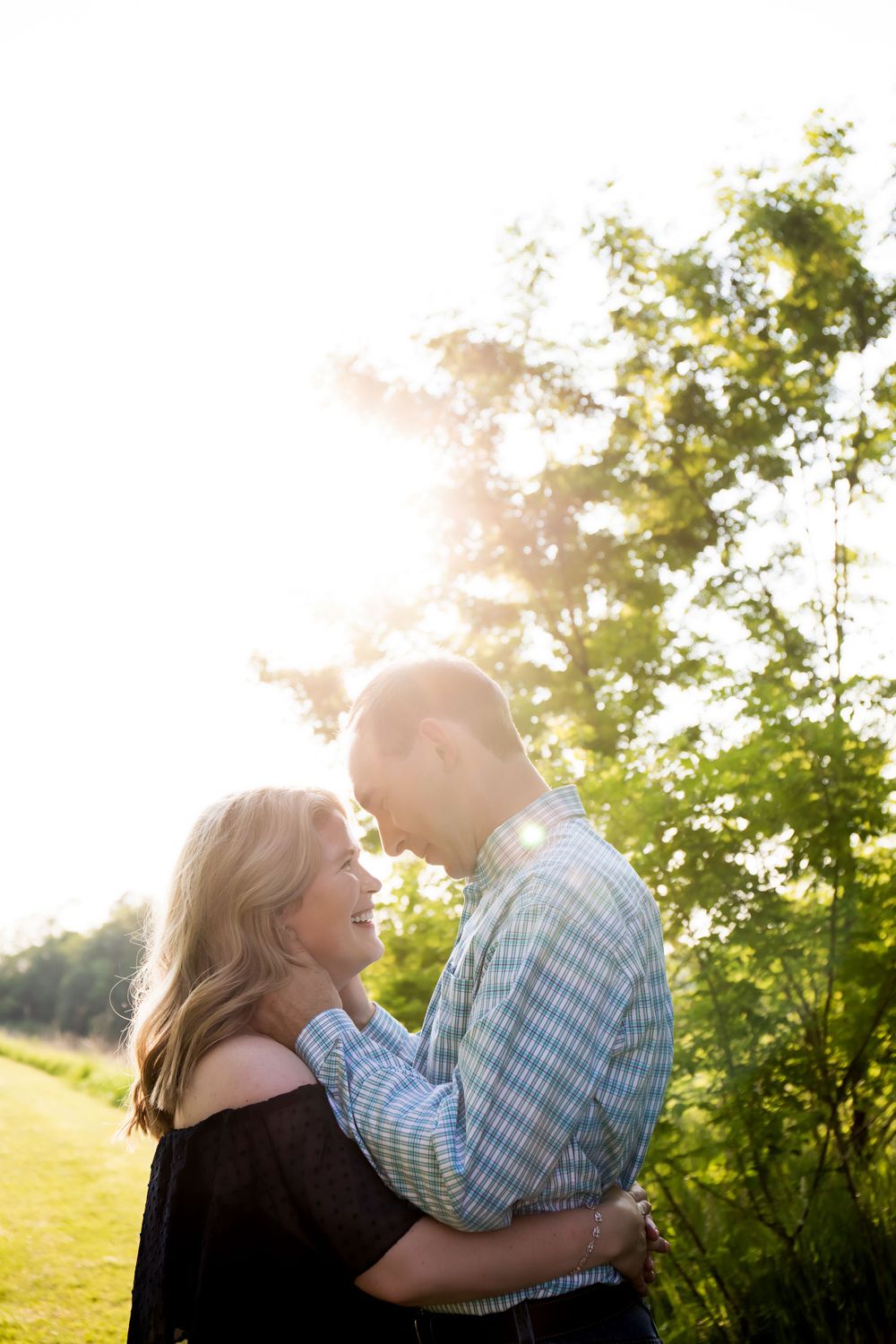 A romantic couple embraces in a sunlit field with trees in the background during golden hour.