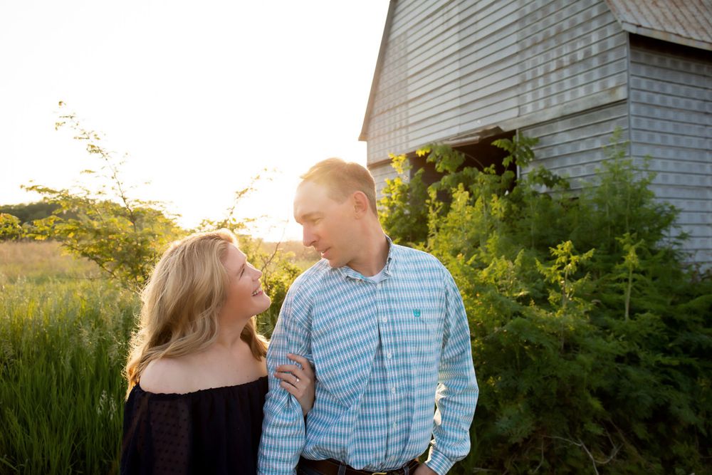A couple gazes at each other lovingly in front of a white barn at sunset.
