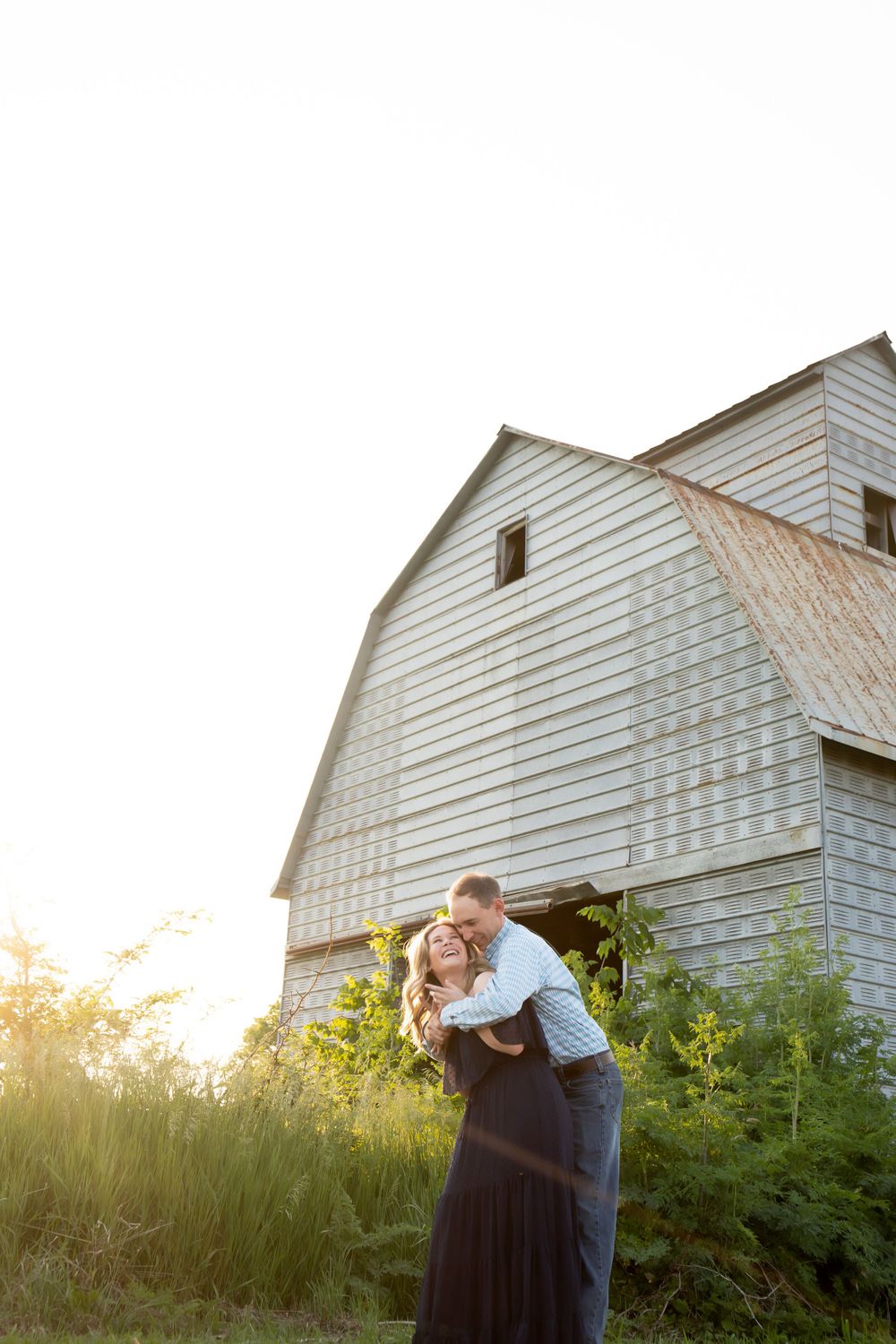 A couple shares a playful moment in front of a weathered white barn at dusk.