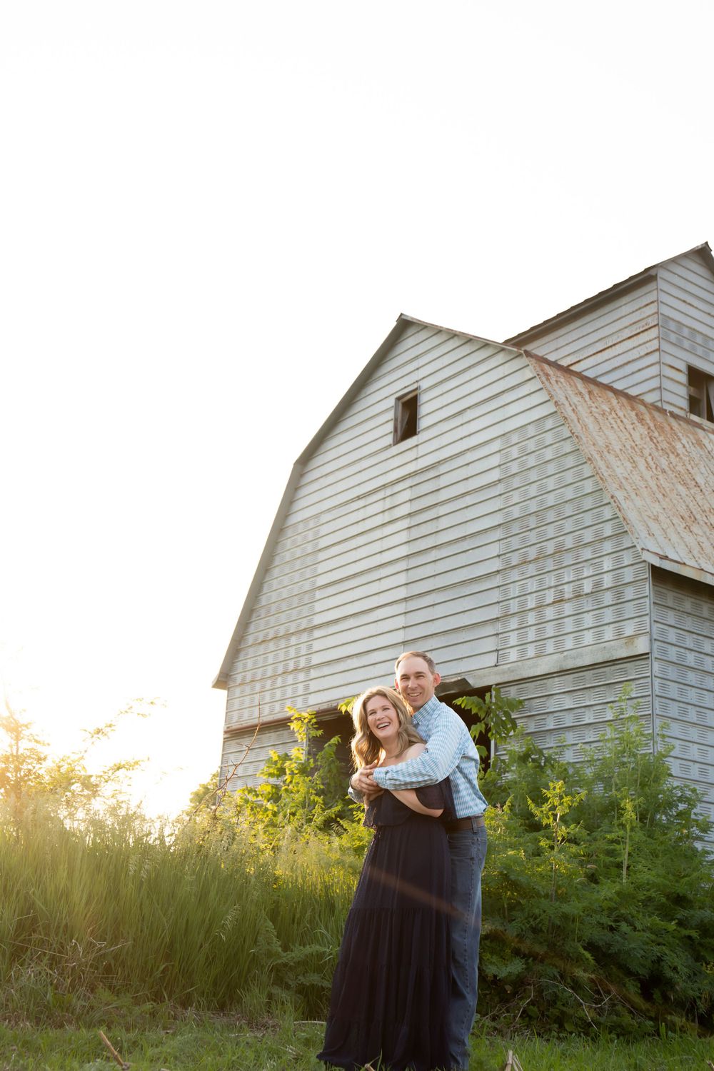 A couple poses together in front of a rustic white barn during sunset.
