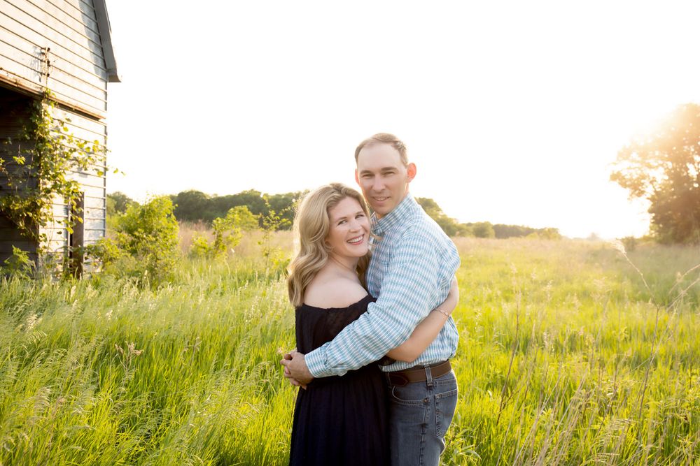 A couple shares a loving embrace in a sunny field next to an old barn.