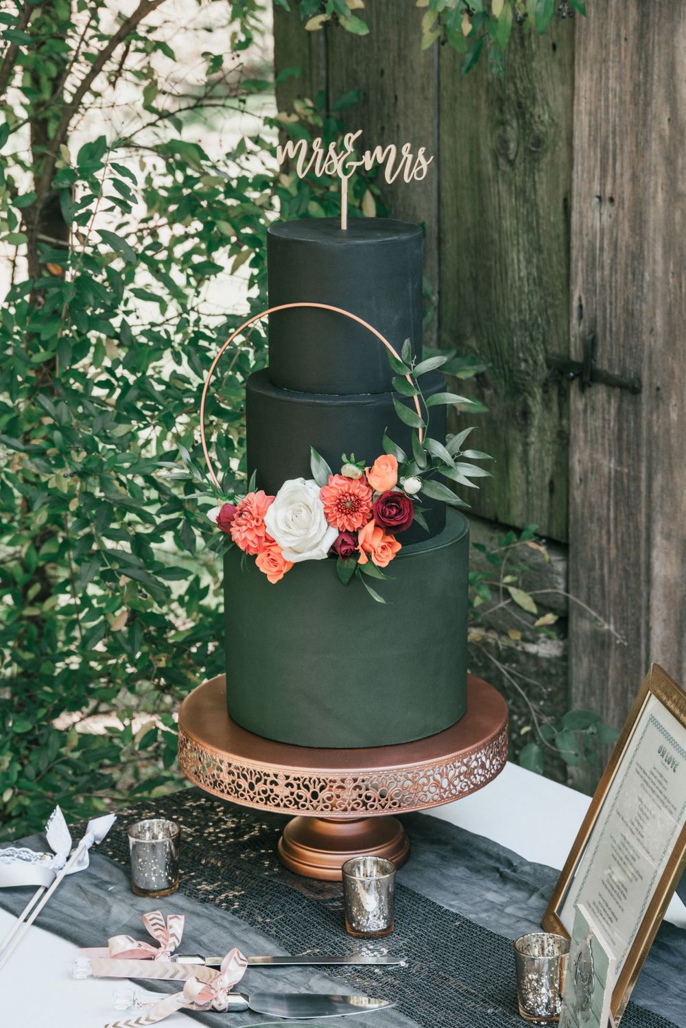 Modern black wedding cake decorated with coral and pink roses on a rose gold stand with Mr & Mrs topper against rustic wood backdrop.