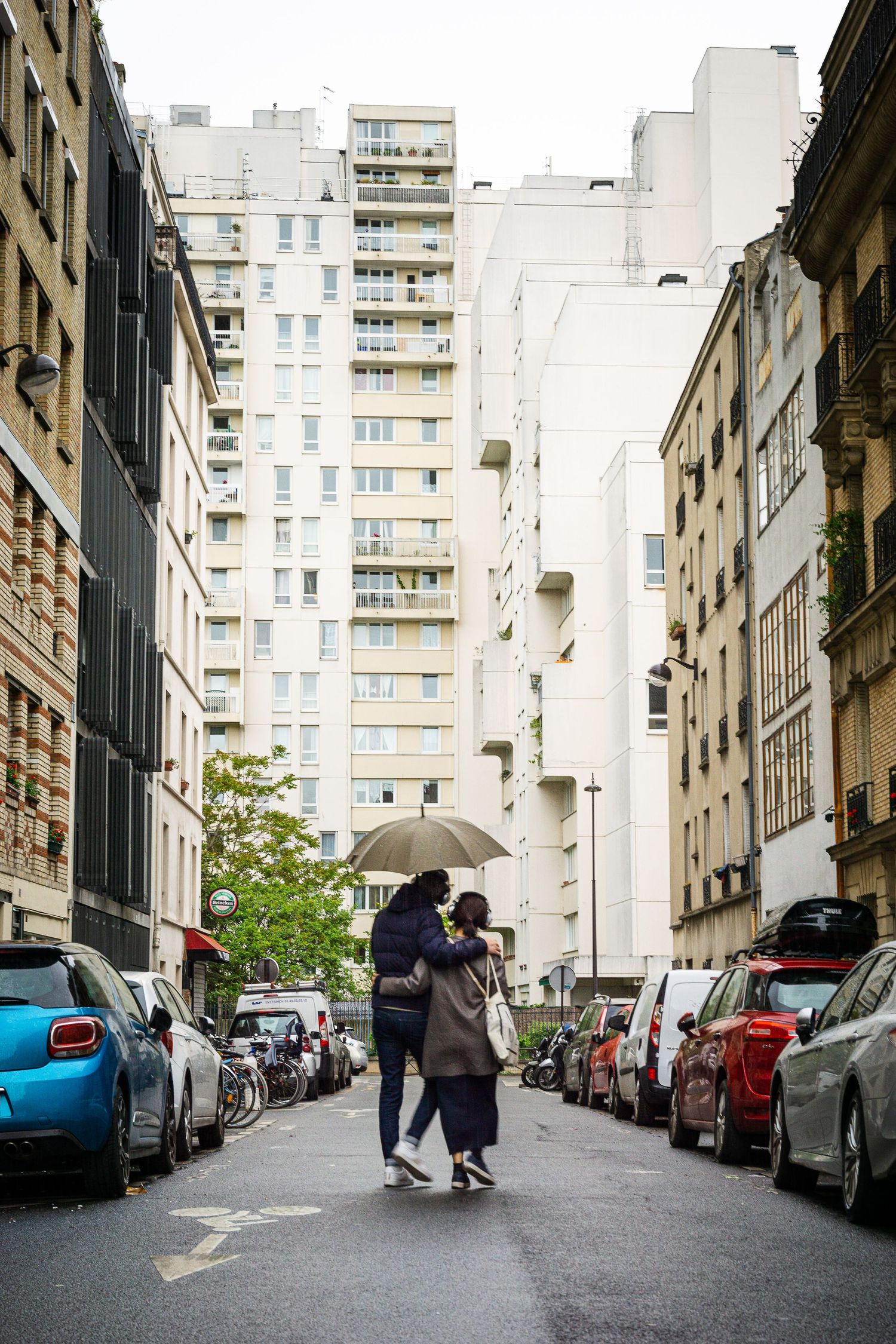 People walking with umbrella on rainy street lined with parked cars and tall buildings.