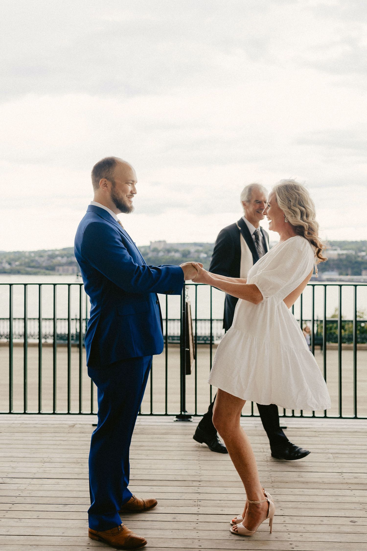 A couple shares an elegant dance moment on a deck overlooking water while an officiant stands nearby.