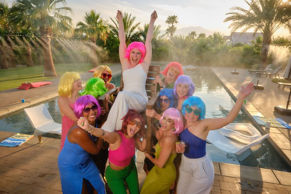 A festive pool party group in bright colors celebrates with raised arms at sunset.