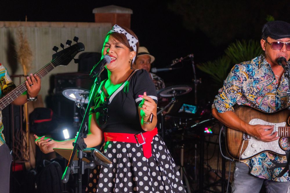 Live band performs on stage with singer in polka dot dress and guitarist in floral shirt.