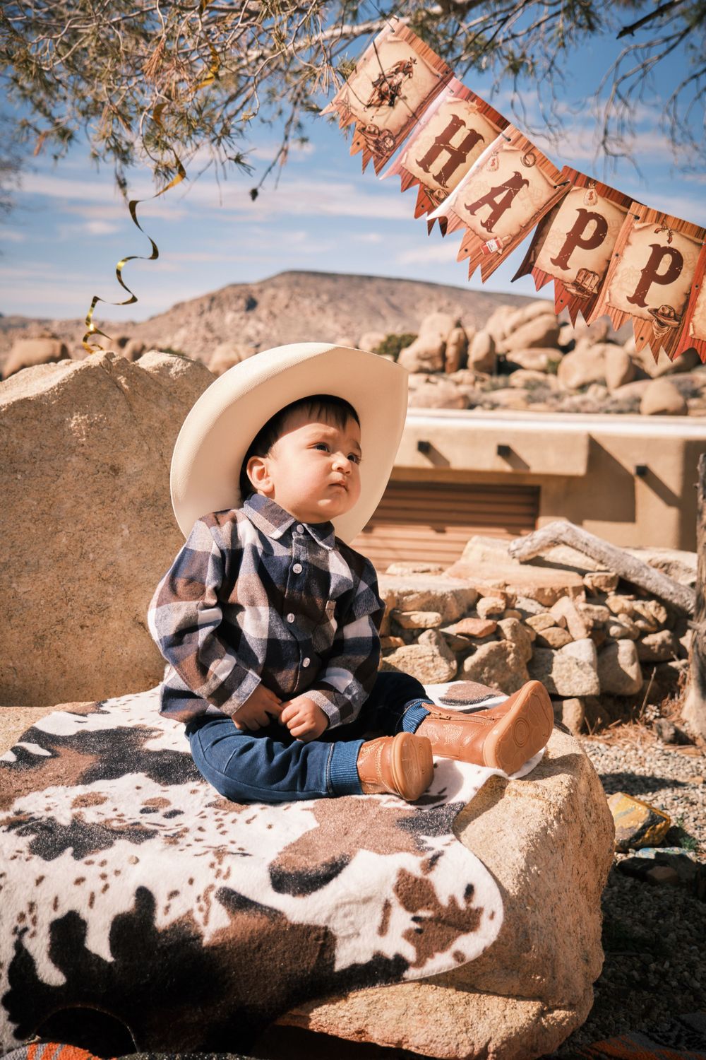 A toddler in western wear sits on a rock with a festive banner in the desert background.