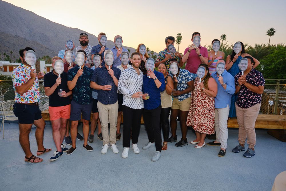 Large group gathering poses together outdoors with mountains visible in the background at sunset.