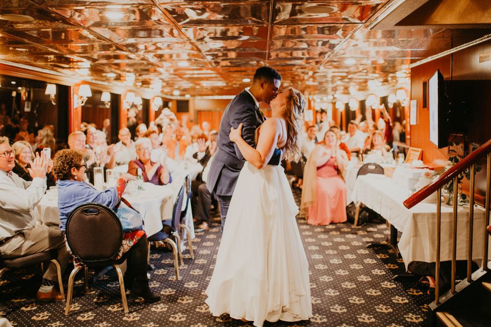 Bride and groom share a first dance kiss surrounded by applauding guests at wedding reception.