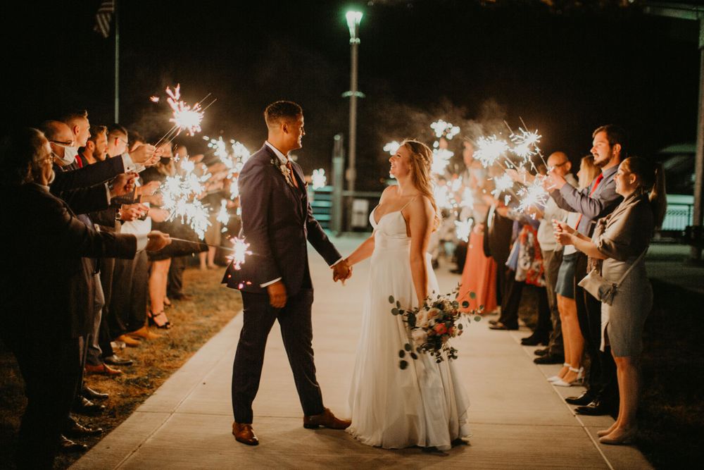 Bride and groom walk through a glowing sparkler exit lined with cheering wedding guests at night.