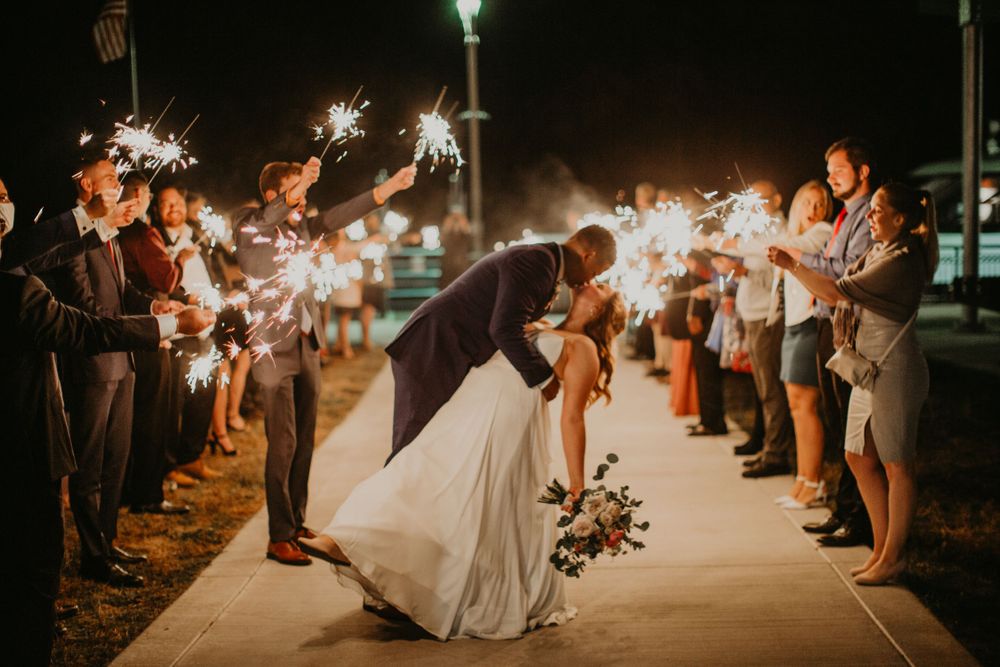 Newlyweds share a dramatic dip kiss during a sparkler exit lined with celebrating wedding guests.