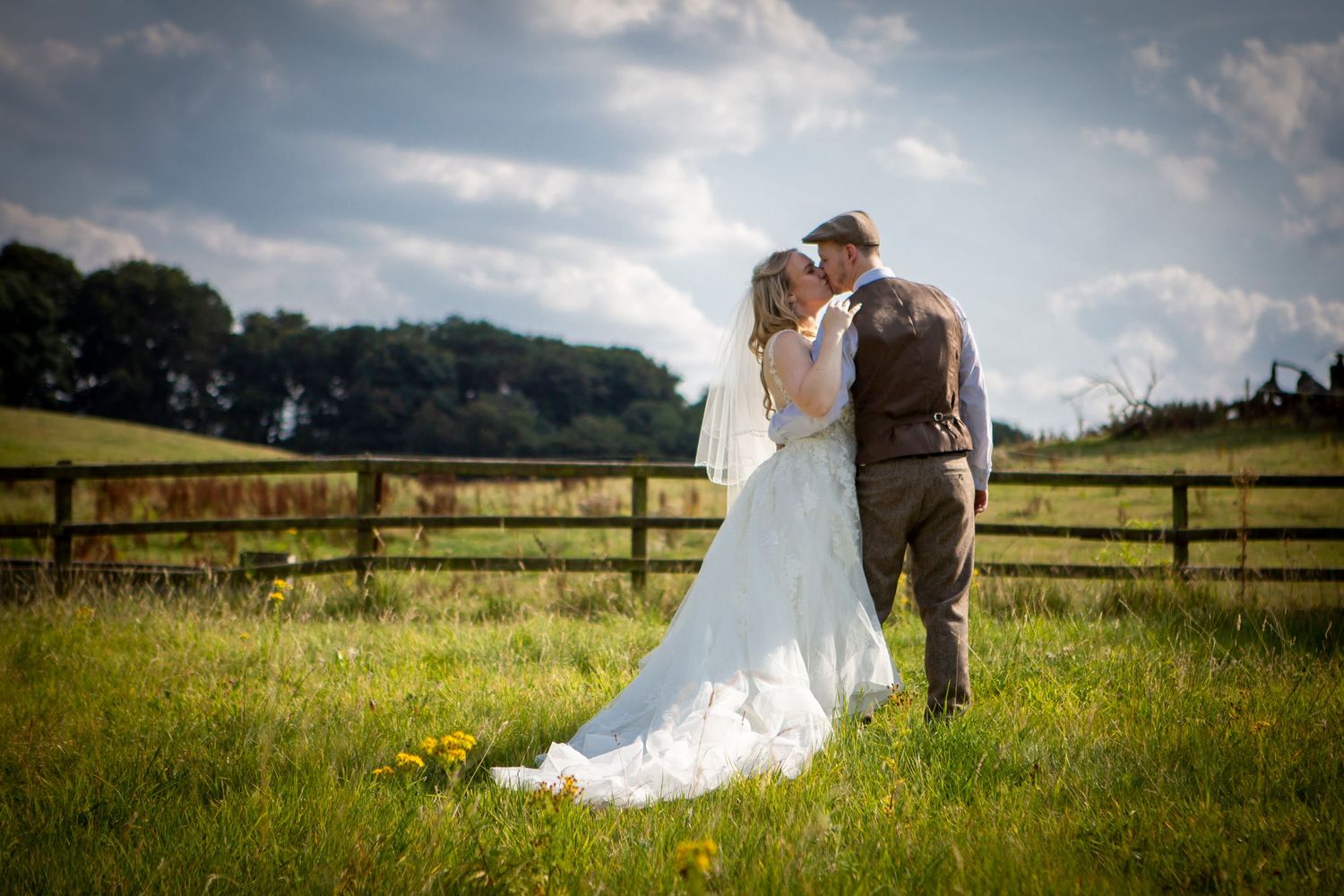 Bride and groom kissing in a sunny green meadow with a wooden fence backdrop.