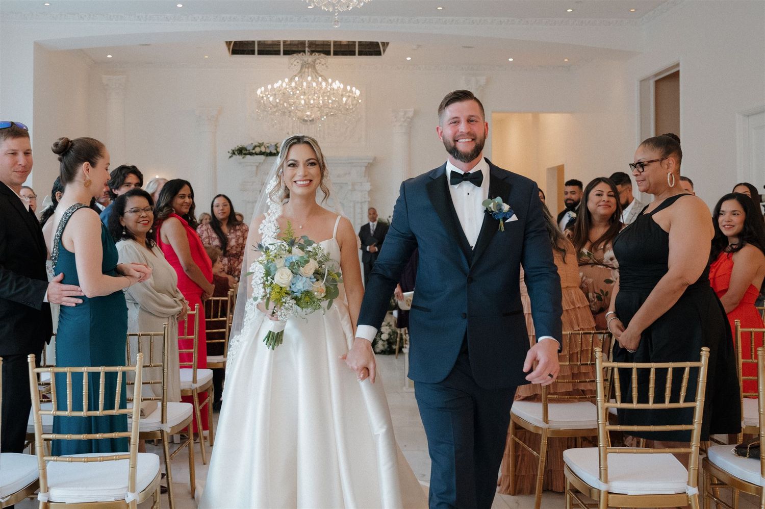 A couple walks down the aisle at their wedding ceremony while guests look on from gold chairs in a bright white venue.