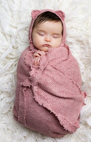 Sleeping baby wearing pink knit bonnet with ears swaddled in matching blanket on white fur.
