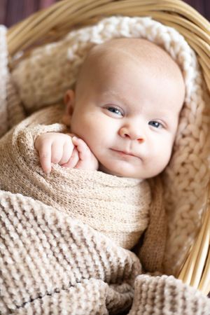 Alert baby wrapped in knit blanket looking up from wicker basket.