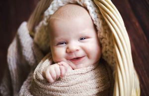 Smiling baby wrapped in knit blanket lying in wicker basket.