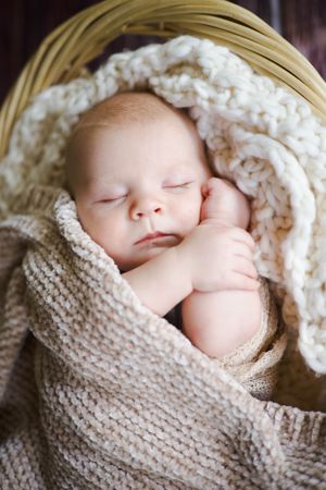 Sleeping newborn baby wrapped in cozy knit blanket in wicker basket.
