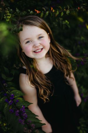 Young child in black dress smiles brightly among purple flowers and green foliage.