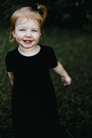 Young child in black dress smiling joyfully outdoors.