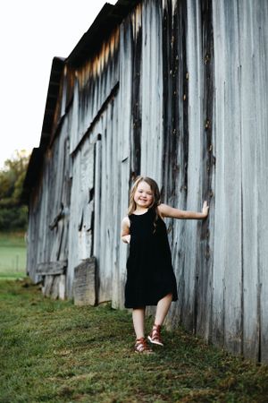 Child in black dress playing by weathered wooden barn wall.