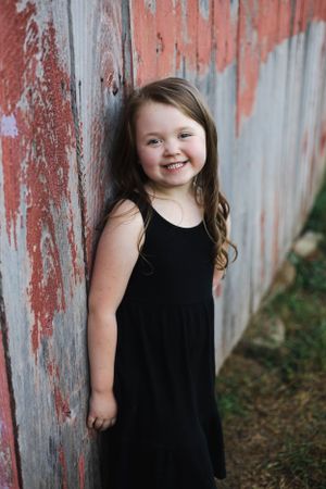 Young person in black dress poses against rustic painted wall.