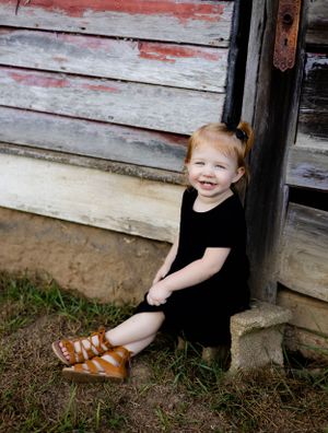 Child in black dress sits against rustic wooden wall wearing brown sandals.