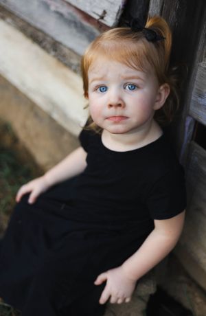 Young child in black dress sitting against rustic wooden wall.
