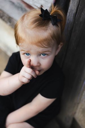 Small child in black with bow peeks shyly at camera against grey wall.