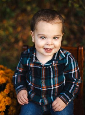 Smiling baby in plaid shirt against fall foliage background.