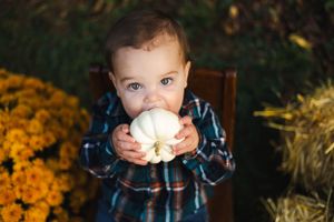 Small child in plaid shirt holds white pumpkin with orange flowers in background.