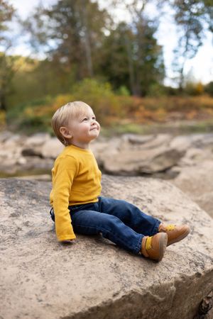 Child in yellow sweater sitting on rock looking up at autumn sky.