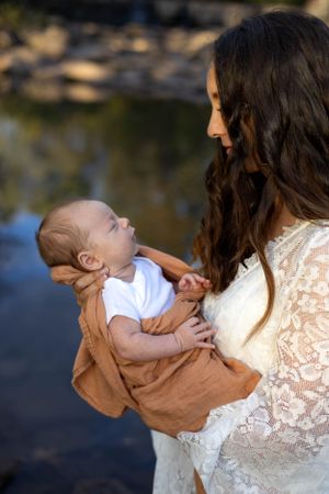 A tender moment between parent and infant beside a lake at sunset, with the baby wearing white and brown.