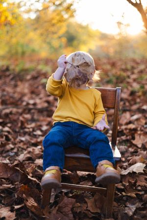 Child in yellow sweater sits on wooden chair among fallen autumn leaves.