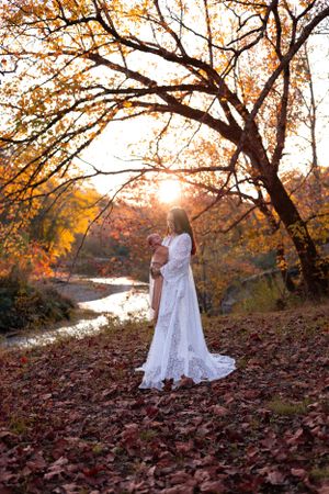 A woman in a flowing white dress holds a baby while standing among autumn leaves at sunset by a stream.