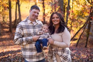 Family wearing coordinated plaid outfits poses together in an autumn forest setting with golden leaves.