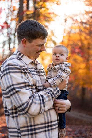 Adult and infant in matching plaid shirts share a tender moment against a backdrop of autumn foliage.