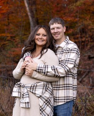 Couple in coordinating plaid outfits embraces against autumn foliage background.