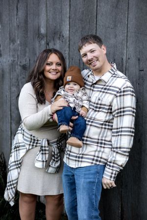 Family poses together against weathered barn wood wearing coordinated plaid and neutral outfits.