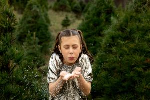 A young girl in a sequined top blows glitter from her hands while standing among Christmas trees.