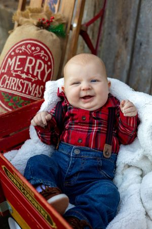 Baby wearing plaid shirt and overalls smiles while sitting in red Christmas sled.