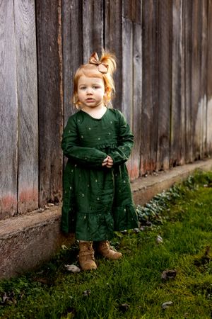 Small child in green dress stands against rustic wooden fence.